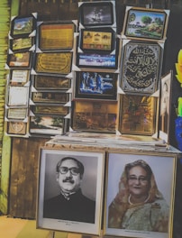 A variety of framed pictures are displayed on a wall, including images of landscapes, calligraphy, and portraits. A stack of frames sits on a table in front, with prominent black-and-white and colored portraits displayed.