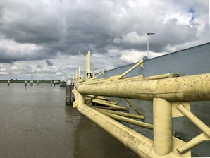 A large, industrial-looking flood barrier stretches across the water with overcast skies looming above. The barrier features substantial yellow metal beams with a weathered appearance. Numerous streetlights line the barrier, and calm water extends into the distance with a green landscape visible on the horizon.