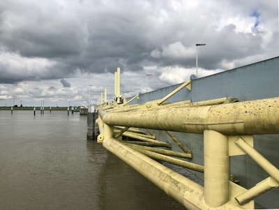 A large, industrial-looking flood barrier stretches across the water with overcast skies looming above. The barrier features substantial yellow metal beams with a weathered appearance. Numerous streetlights line the barrier, and calm water extends into the distance with a green landscape visible on the horizon.