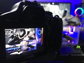 A digital camera is positioned to capture an image displayed on its screen. The screen shows a vivid and colorful scene featuring a pair of Nike sneakers, vibrant neon lights, and graphic elements in the background. The setup suggests a creative and modern product photography shoot.