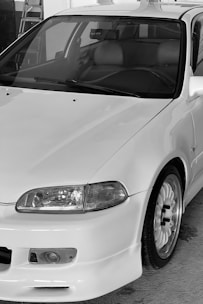 A close-up view of a white car with a sleek, aerodynamic design. Key features include a prominent front bumper, rounded headlights, and a detailed wheel with a sporty rim. The interior appears to have leather seats, visible through the front windshield.