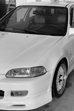 A close-up view of a white car with a sleek, aerodynamic design. Key features include a prominent front bumper, rounded headlights, and a detailed wheel with a sporty rim. The interior appears to have leather seats, visible through the front windshield.