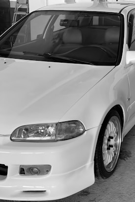 A close-up view of a white car with a sleek, aerodynamic design. Key features include a prominent front bumper, rounded headlights, and a detailed wheel with a sporty rim. The interior appears to have leather seats, visible through the front windshield.