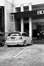 A black and white image featuring a parked white Honda CR-V with customized wheels in front of a building with a prominent BDO sign. A person is seen entering the building through glass doors near a flight of stairs.