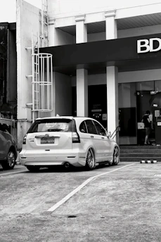 A black and white image featuring a parked white Honda CR-V with customized wheels in front of a building with a prominent BDO sign. A person is seen entering the building through glass doors near a flight of stairs.