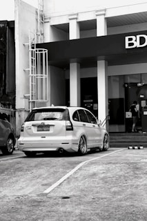 A black and white image featuring a parked white Honda CR-V with customized wheels in front of a building with a prominent BDO sign. A person is seen entering the building through glass doors near a flight of stairs.