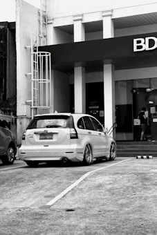 A black and white image featuring a parked white Honda CR-V with customized wheels in front of a building with a prominent BDO sign. A person is seen entering the building through glass doors near a flight of stairs.