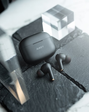 A pair of black wireless earbuds and their charging case are positioned on a textured, dark slate surface. Nearby, a transparent glass cube and a prism create subtle reflections, adding a modern, minimalist aesthetic.