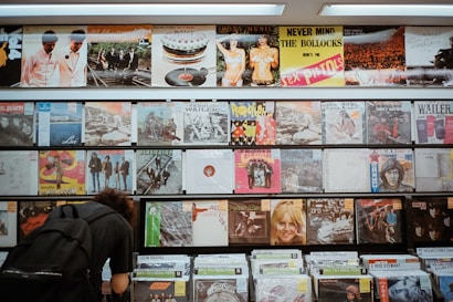 A wall display of vinyl records featuring a variety of album covers, including notable bands and artists. The albums are arranged in multiple rows on shelves with a mixture of classic rock and other genres. A person with curly hair and a backpack is browsing through the lower section of the records.