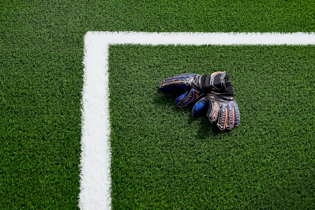 A pair of colorful goalkeeper gloves lies on a green soccer field near the white boundary line.