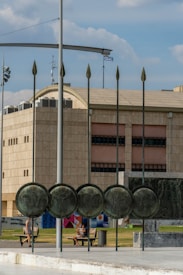 A large, modern building with a beige facade dominates the background. In front of it is a sculpture featuring several large round shields with intricate designs. Metal poles with spear-like tips extend upward from the sculpture. A Greek flag flies in the background. Two people are sitting on a bench in a small park area, which also has a colorful mural.