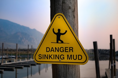 A yellow triangular warning sign is mounted on a wooden post. The sign features a black silhouette of a person with raised arms, standing on a horizontal line that suggests instability, with the words 'Danger Sinking Mud' below the figure. The background includes a partially visible landscape with mountains and water, possibly a dock or lake area.
