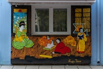 A mural featuring stylized figures in traditional Asian attire engaged in various activities. The vibrant colors and illustrative style suggest cultural or educational themes. One figure in a green outfit sits with a contemplative expression, while another in red kneels, possibly writing or reading from a scroll. A third figure in yellow holds a book. Decorative backdrops of windows and patterns add depth to the scene.
