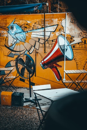 A vibrant mural featuring a stylized bird alongside a large red and white megaphone is painted on an orange wall. In front of the mural, there is a black stand fan and several foldable chairs and tables set up on a pebbled ground.