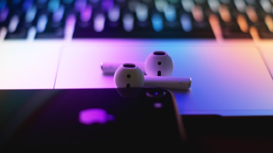 A close-up of wireless earbuds resting on a laptop with illuminated keys. The scene is bathed in a gradient of colorful lights, creating a sleek and modern aesthetic. The focus includes a partially visible smartphone placed nearby.