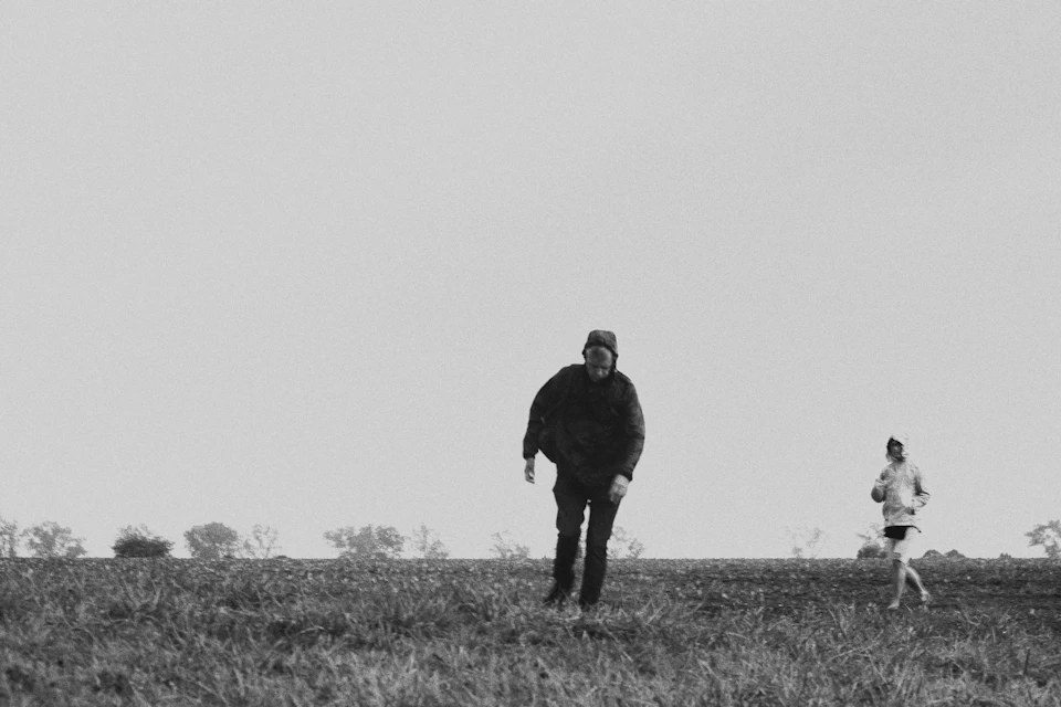 Man and woman walk through a field with trees and bushes behind them together