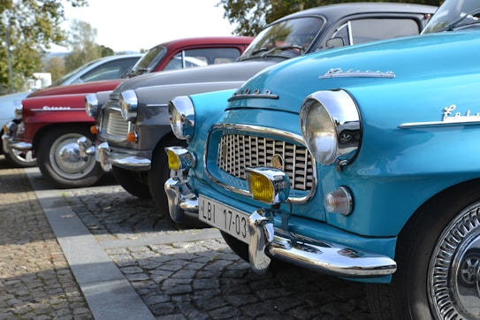 A row of vintage cars parked on a cobblestone street, featuring models in blue, silver, and red. The cars have distinct chrome accents and classic designs, indicating they are from the mid-20th century.