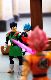 Three Dragon Ball Z action figures are positioned in a dynamic setting. The figure in the foreground is dressed in an orange gi with pink hair, facing away from the camera. Another figure wearing a green and black outfit with a red cape is focused and seems to be looking at the pink-haired character. The third figure, in the background, has blue hair and is wearing a blue and black outfit. There is a pink light effect from the orange gi figure, adding emphasis.