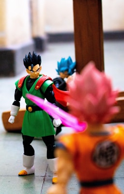 Three Dragon Ball Z action figures are positioned in a dynamic setting. The figure in the foreground is dressed in an orange gi with pink hair, facing away from the camera. Another figure wearing a green and black outfit with a red cape is focused and seems to be looking at the pink-haired character. The third figure, in the background, has blue hair and is wearing a blue and black outfit. There is a pink light effect from the orange gi figure, adding emphasis.