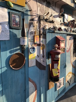 A wall filled with eclectic decorations including various clocks, frames, a red postbox, and eclectic artwork. The setting conveys a whimsical, artistic vibe with mixed media and unique objects fixed on textured walls under warm lighting.