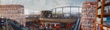 A large, modern library with towering cylindrical bookshelves filled with books. The space is bustling with people, some reading and others walking around. An expansive glass ceiling lets in natural light, enhancing the open, airy atmosphere. There are multiple levels accessible by escalators.