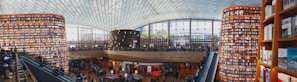 A large, modern library with towering cylindrical bookshelves filled with books. The space is bustling with people, some reading and others walking around. An expansive glass ceiling lets in natural light, enhancing the open, airy atmosphere. There are multiple levels accessible by escalators.