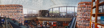 A large, modern library with towering cylindrical bookshelves filled with books. The space is bustling with people, some reading and others walking around. An expansive glass ceiling lets in natural light, enhancing the open, airy atmosphere. There are multiple levels accessible by escalators.