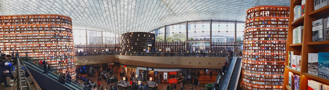 A large, modern library with towering cylindrical bookshelves filled with books. The space is bustling with people, some reading and others walking around. An expansive glass ceiling lets in natural light, enhancing the open, airy atmosphere. There are multiple levels accessible by escalators.