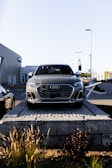 A silver Audi SUV is displayed prominently at the forefront, elevated on a stone platform in front of a car dealership. The building and additional vehicles are visible in the background, with clear skies overhead.