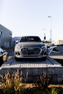 A silver Audi SUV is displayed prominently at the forefront, elevated on a stone platform in front of a car dealership. The building and additional vehicles are visible in the background, with clear skies overhead.