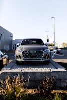 A silver Audi SUV is displayed prominently at the forefront, elevated on a stone platform in front of a car dealership. The building and additional vehicles are visible in the background, with clear skies overhead.