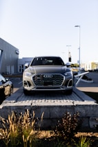 A silver Audi SUV is displayed prominently at the forefront, elevated on a stone platform in front of a car dealership. The building and additional vehicles are visible in the background, with clear skies overhead.