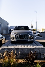 A silver Audi SUV is displayed prominently at the forefront, elevated on a stone platform in front of a car dealership. The building and additional vehicles are visible in the background, with clear skies overhead.