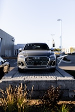 A silver Audi SUV is displayed prominently at the forefront, elevated on a stone platform in front of a car dealership. The building and additional vehicles are visible in the background, with clear skies overhead.