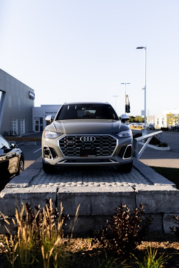 A silver Audi SUV is displayed prominently at the forefront, elevated on a stone platform in front of a car dealership. The building and additional vehicles are visible in the background, with clear skies overhead.