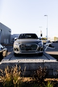 A silver Audi SUV is displayed prominently at the forefront, elevated on a stone platform in front of a car dealership. The building and additional vehicles are visible in the background, with clear skies overhead.