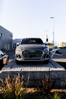 A silver Audi SUV is displayed prominently at the forefront, elevated on a stone platform in front of a car dealership. The building and additional vehicles are visible in the background, with clear skies overhead.