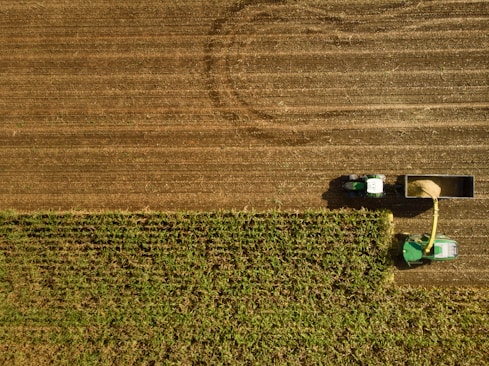 An aerial view of a farmland scene with a tractor and a harvester working on a field. The landscape is divided into two main sections: the top has harvested brown, bare earth, while the bottom is lush with green crops. The harvester is connected to a trailer, collecting the harvested material.