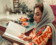 A woman wearing a colorful outfit and headscarf is sitting on a couch, holding an open book with text in a foreign language. In front of her, there is a table with various fruits, sweets, and a vase with flowers. The room is decorated with plants and patterned wallpaper, creating a cozy and inviting atmosphere.