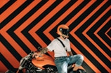 A person wearing an orange motorcycle helmet sits on an orange Ducati motorcycle. The background features a bold geometric pattern with alternating orange and black chevrons, creating a striking visual impact. The person is dressed casually in a white shirt and jeans, with a tattoo visible on the arm.