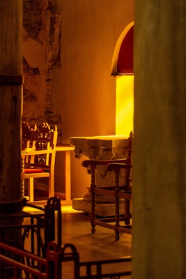 The image depicts an interior scene with a warm, golden lighting. It features intricately designed wooden chairs arranged around a stone table or altar. The walls and the floor show texture typical of aged, possibly historic structures. The overall setting has an antique and rustic charm, enhanced by the glowing illumination.