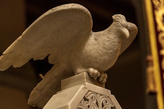 A detailed stone sculpture of a bird, possibly an eagle, with elaborately carved feathers is perched on a decorative pedestal. The sculpture exhibits intricate patterns and appears to be part of a classical or historical decor.