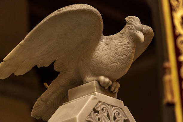 A detailed stone sculpture of a bird, possibly an eagle, with elaborately carved feathers is perched on a decorative pedestal. The sculpture exhibits intricate patterns and appears to be part of a classical or historical decor.