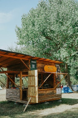 A rustic wooden cabin serves as an outdoor kiosk, set within a green, leafy environment. The structure is made of logs with a pitched roof and open front, displaying various goods for sale. A large tree provides shade behind the cabin, while a few tents are visible in the background, indicating a camping area.