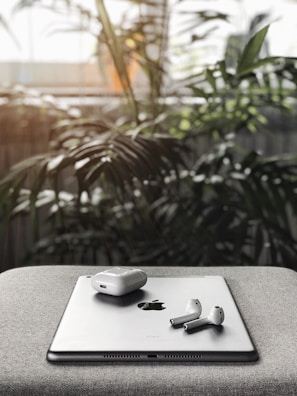 A pair of wireless earbuds and their charging case rest on a tablet with an Apple logo, placed on a gray fabric surface. In the background, large green indoor plants are softly blurred with sunlight filtering through.