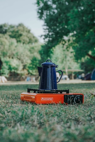A blue enamel coffee pot is placed on an orange portable camping stove set on grassy ground. The background consists of blurred green trees and a few tents, suggesting a camping environment.