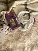 A cozy arrangement featuring a fluffy blanket with a plaid pattern, a white bib with a purple floral design, and a package of lavender sachets adorned with an image of a teddy bear. Additionally, there are soft toy animals arranged nearby, including a bear and a rabbit.