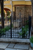 A stone tomb surrounded by intricate black metal fencing is situated in a serene outdoor setting. Lush green plants grow around the base of the tomb. The background features an old stone wall and an archway with classical architectural elements such as columns and arches. The area is paved with stone tiles, and trees provide shade.