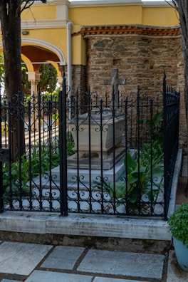 A stone tomb surrounded by intricate black metal fencing is situated in a serene outdoor setting. Lush green plants grow around the base of the tomb. The background features an old stone wall and an archway with classical architectural elements such as columns and arches. The area is paved with stone tiles, and trees provide shade.