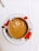A porcelain cup filled with milk tea sits on a matching saucer featuring red floral designs. A splash of milk is captured mid-air, creating ripples and bubbles in the tea. The setting is on a white tablecloth.